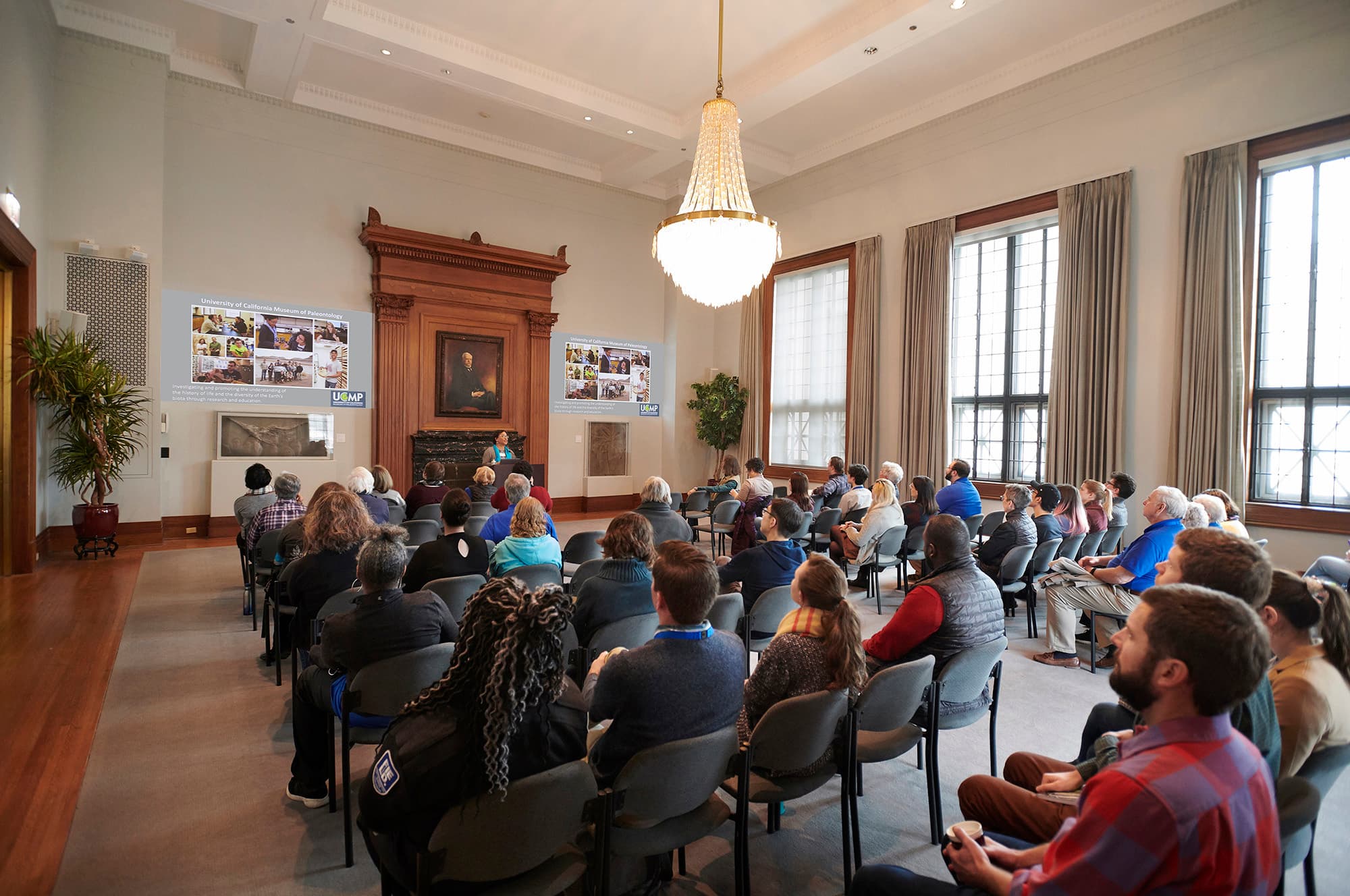 A group of people gather in a room, seated, to listen to a lecture. The speaker stands behind a podium with images projected on the wall behind them.