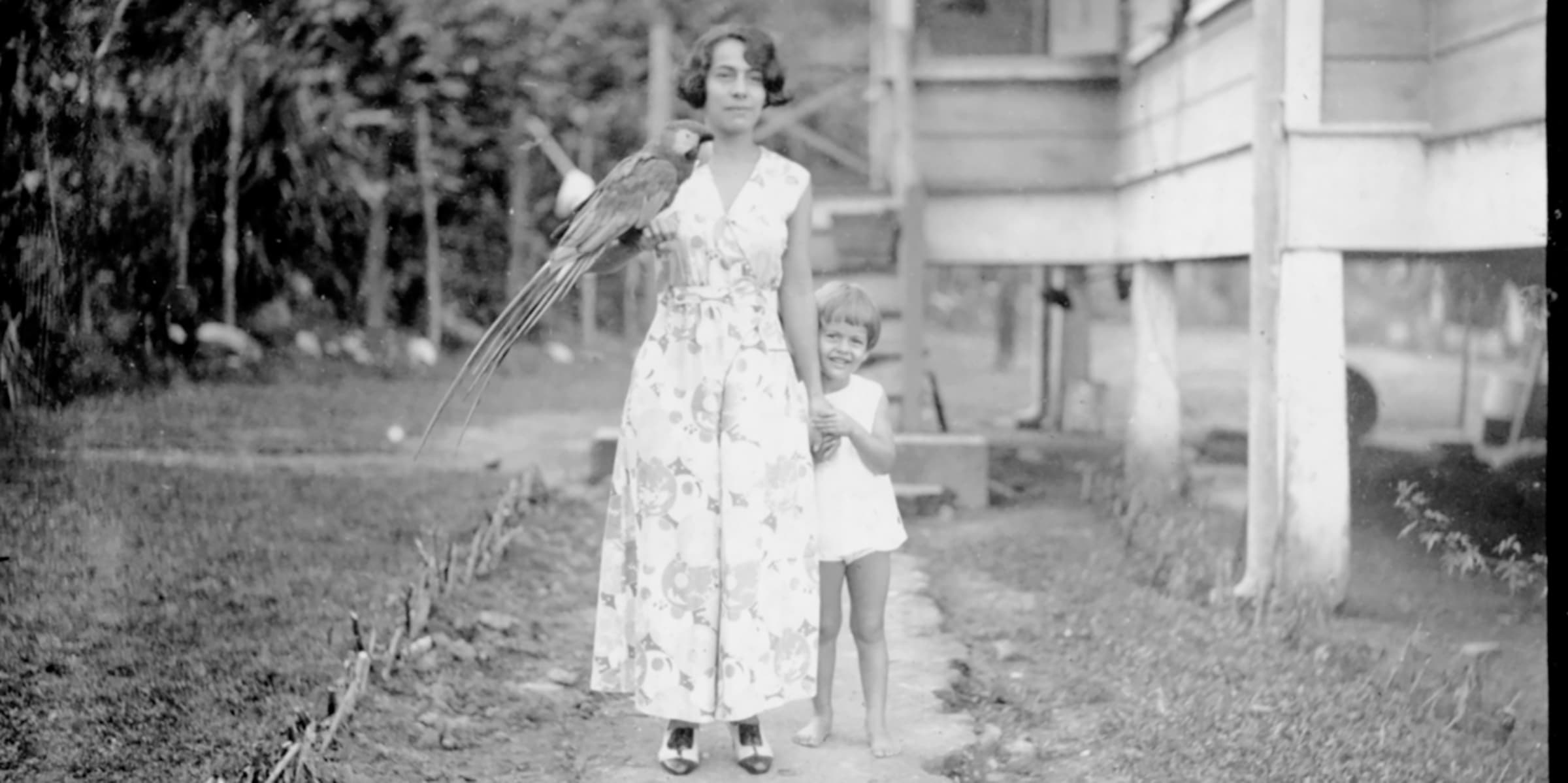 Black and white photograph of a woman in a floral dress with a large bird on her hand, and a small child smiling and standing at her side