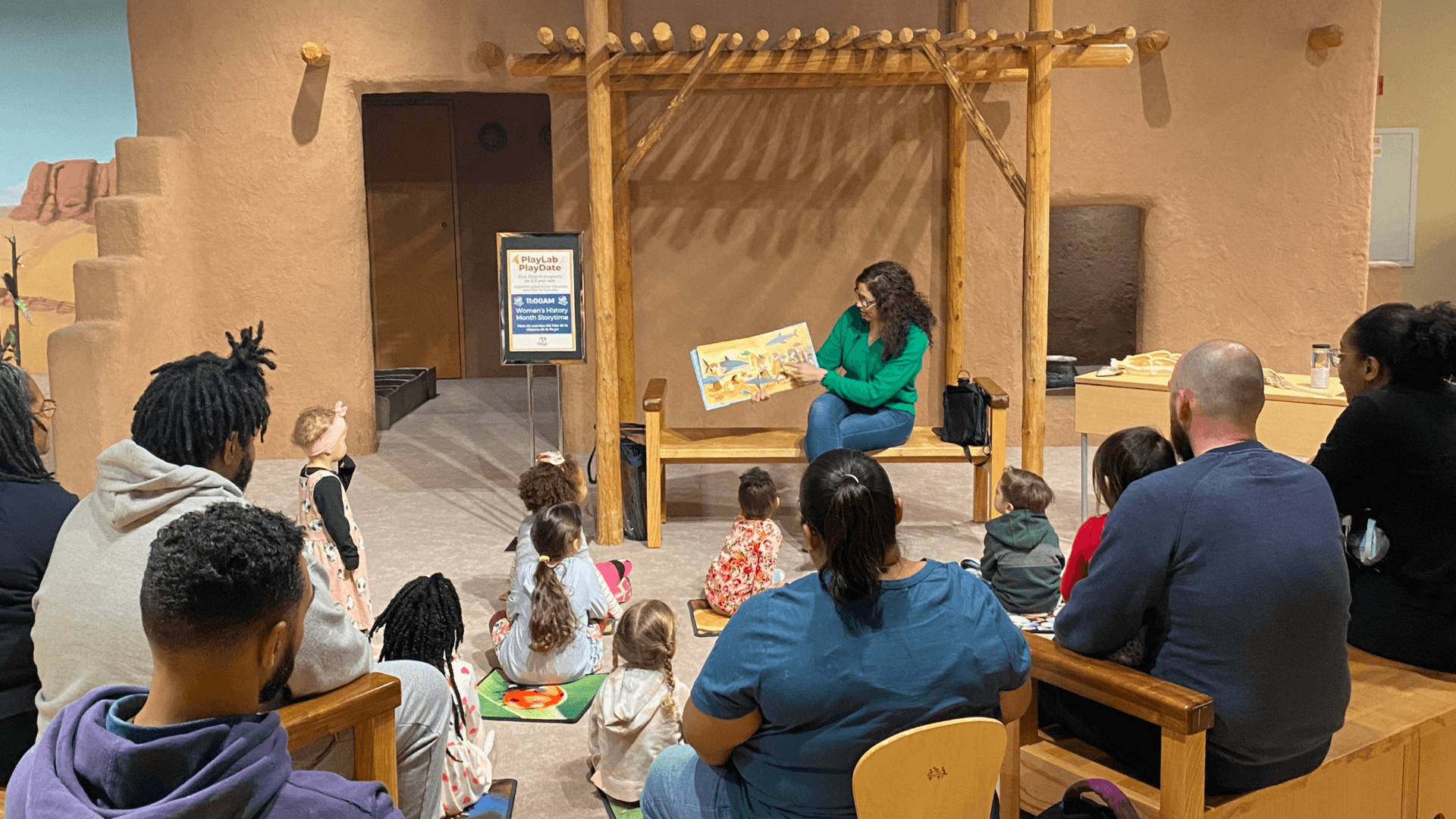 a woman reads a story to a group of young children sitting on the floor in front of her. Several adults are seated behind the children.